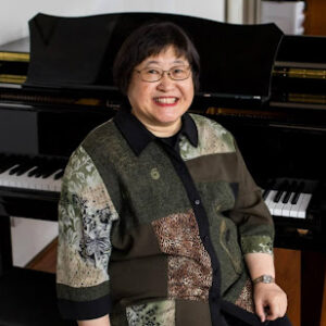 Chen Yi, composer, sitting at a piano, facing the camera and smiling