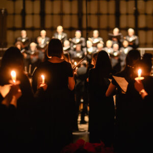 Photo of silhouetted treble singers holding candles, with tenors and basses in the distance in a group.